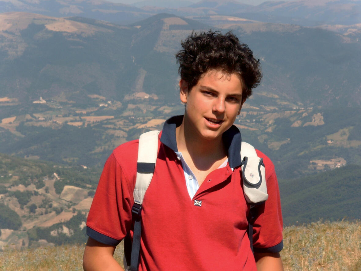 Carlo Acutis mit dunklem Haar in rotem Shirt, vor einer hügeligen Landschaft in Italien.