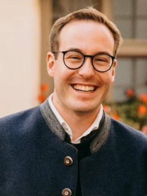Alexander Grotz mit Brille und Trachtenjacke vor blühendem Hintergrund.